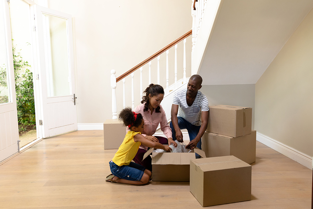 Front view of a mixed race couple and their young daughter kneeling in the hallway of their new home unpacking boxes together on the floor